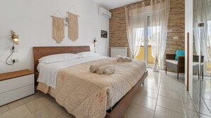 Un dormitorio con cama de madera, ropa de cama blanca, una mesita de noche, aire acondicionado instalado en la pared, una ventana con cortinas translúcidas y vistas al exterior.