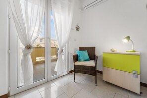 Ein Zimmer mit einer Schiebetür aus Glas, ein Korbstuhl mit Kissen, eine grüne und weiße Kommode sowie eine Schreibtischlampe.