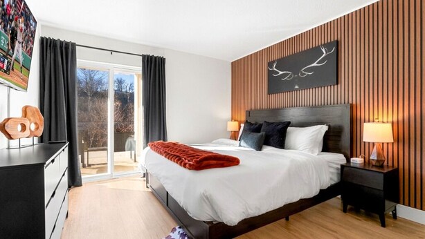 Een moderne slaapkamer met een groot bed, een houten hoofdbord, een kunstwerk aan de muur en een glazen schuifdeur naar een buitenruimte.
