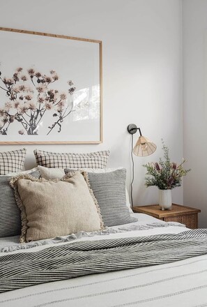 Et soveværelse med seng, puder, et natbord med lampe, et indrammet kunstværk og en vase med blomster.