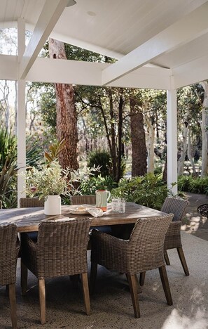 Eine Terrasse im Freien mit einem runden Tisch und Korbstühlen, umgeben von Grünpflanzen und Bäumen.
