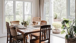 A dining area with a wooden table set for six, surrounded by wicker chairs, with large windows offering a view of greenery outside. - Scribbly Gums Berrara (Berrara)