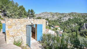 Ein Steinhaus mit einer Holzdielen-Terrasse, einer blauen Tür und Blick auf eine gebirgige Landschaft.