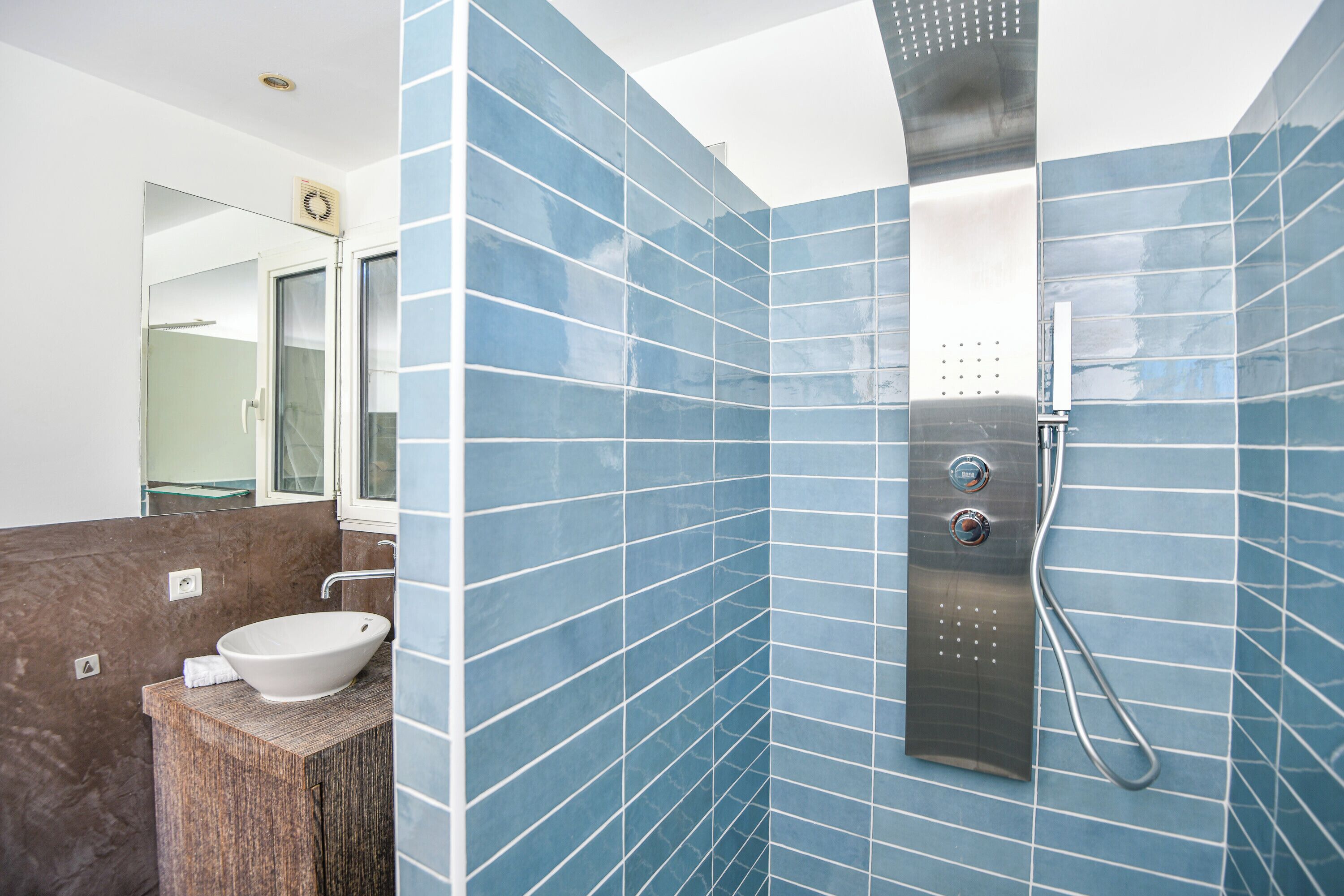 Un bagno con un lavandino bianco, uno specchio, una doccia con soffione a mano e pareti rivestite di piastrelle blu.