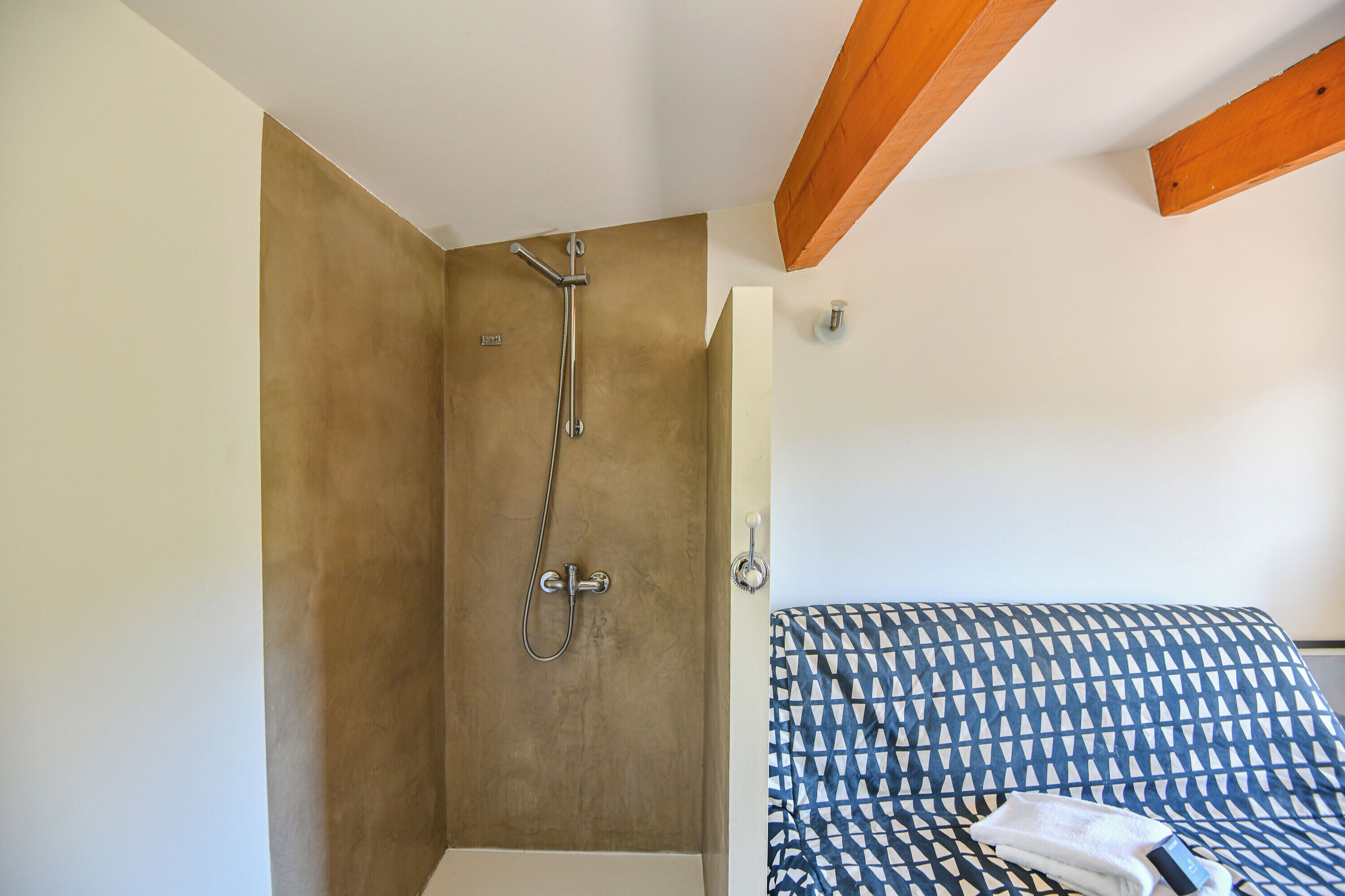 A shower with a handheld showerhead, a towel on the floor, and a bed with a patterned bedspread.