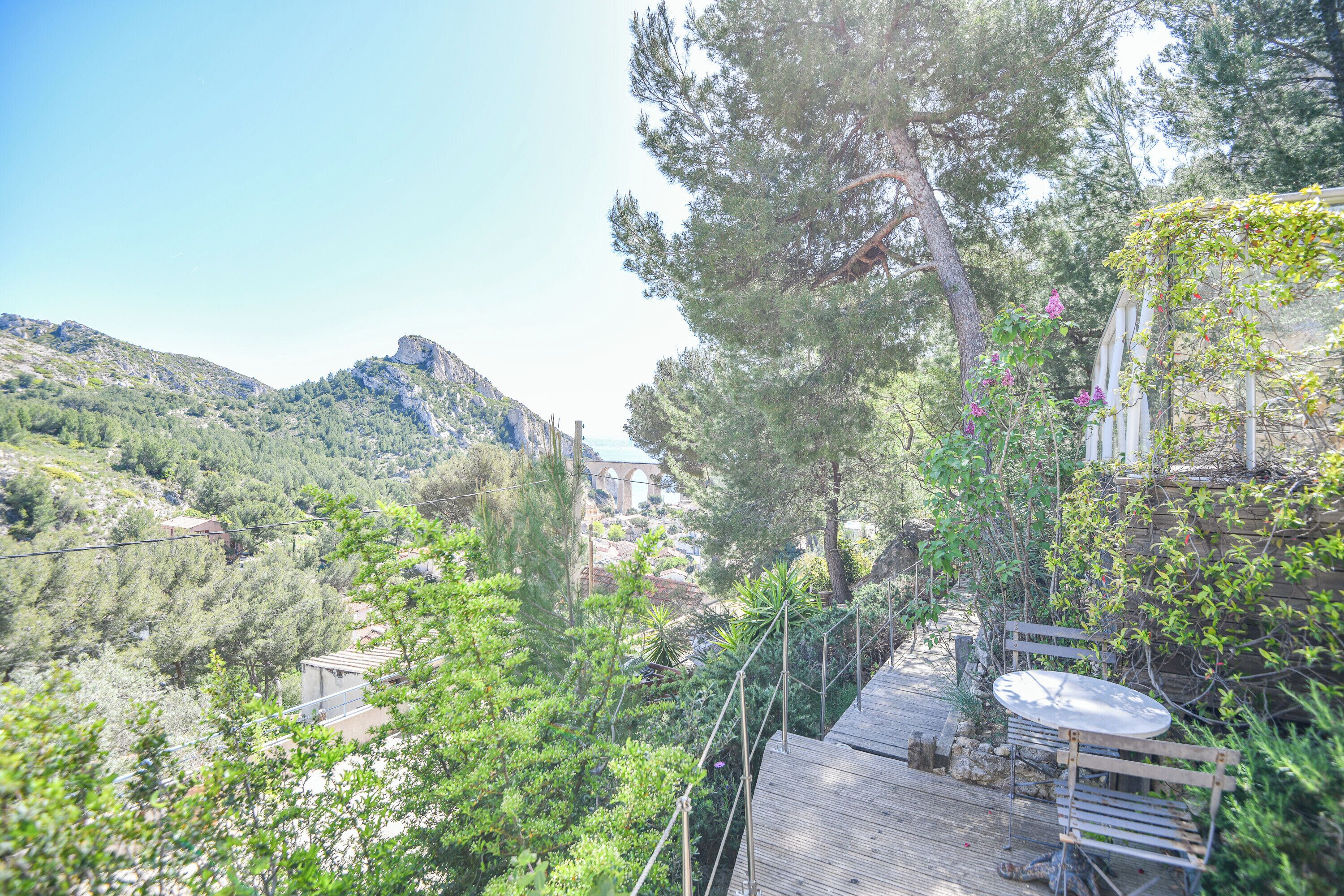 Una terraza de madera con una mesa y banco, rodeada de vegetación y con vistas a un paisaje montañoso.
