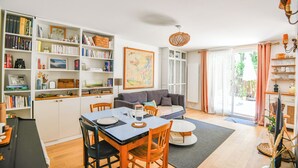 Et godt oplyst opholdsstue med spiseplads, en reol, en sofa og et stort vindue.
