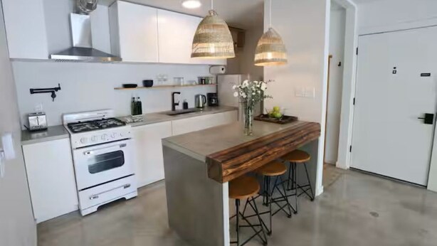 Cozinha moderna com armários brancos, fogão embutido, luminárias pendentes e ilha de madeira com banquetas.