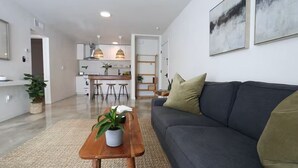 Uma sala de estar moderna com um sofá, uma mesa de centro com uma planta e uma cozinha ao fundo.