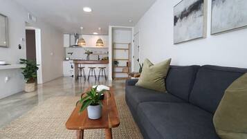 A modern living room with a sofa, a coffee table with a plant, and a kitchen in the background.