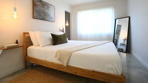 Quarto com cama de madeira, roupa de cama branca, mesa de cabeceira, luminária na parede, um quadro e janela com cortinas.