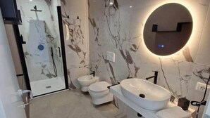 A modern bathroom with a marble wall, a round mirror, a white sink, and a shower area.