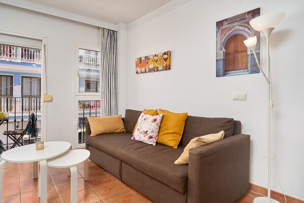 Un salon avec un canapé marron, des tables rondes blanches, un lampadaire, un mur orné d’une œuvre d’art colorée et un balcon offrant une vue sur les bâtiments.
