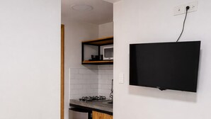 Una cocina compacta con microondas, estufa y refrigerador, un televisor montado en la pared y una cama con ropa blanca.