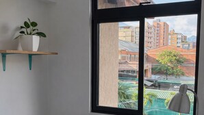 Una habitación con cama, ventana con vistas a edificios, una planta en un estante y una lámpara.