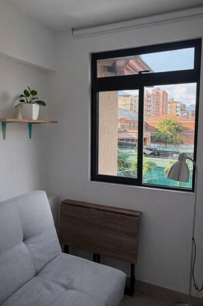 A room with a bed, a window with a view of buildings, a plant on a shelf, and a lamp. - Cozy loft in Laureles (Medellín)