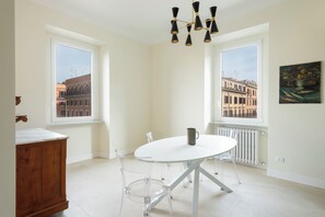 Ein Zimmer mit einem weißen Tisch, transparenten Stühlen, einer Holzkommode, einer schwarzen Pendelleuchte und einem Gemälde an der Wand.