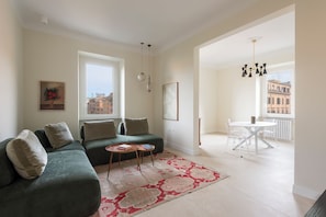 Ein Wohnzimmer mit einem grünen Sofa, einem kleinen runden Tisch, einem gemusterten Teppich, einem Essbereich mit weißem Tisch und Stühlen sowie Blick auf Gebäude durch große Fenster.