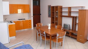 Een compacte keuken met oranje kastjes, een eettafel met stoelen, een televisie en een boekenplank.