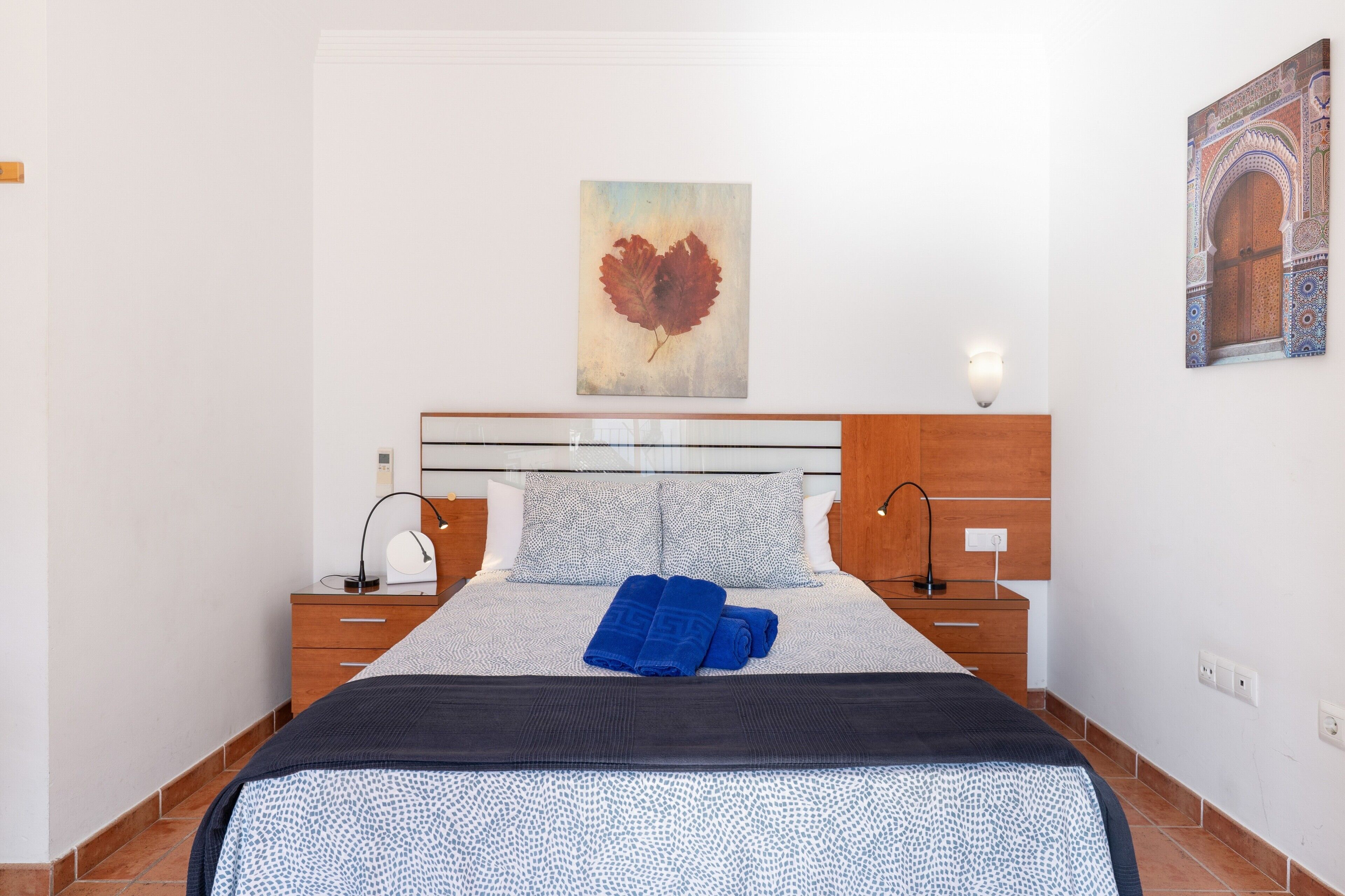A neatly made bed with a patterned bedspread, two bedside tables with lamps, a wall-mounted artwork, and a decorative wall panel.