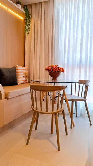 Ein modernes Wohnzimmer mit Glastisch, Holzstühlen, einer Couch mit Kissen und einem großen Fenster mit transparenten Vorhängen.