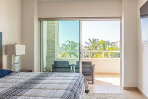 Une chambre avec un lit, une table de chevet, une lampe, un balcon avec des chaises et une vue sur des palmiers et des immeubles.