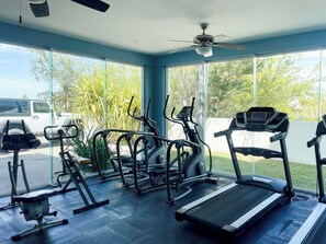 Ein Fitnessstudio mit Trainingsgeräten, Laufband und großen Fenstern mit Blick auf Bäume und ein parkendes Fahrzeug.