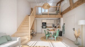 Een loft-achtige kamer met een houten trap, een eethoek met groene stoelen, een keuken met moderne apparatuur, en een groot raam.