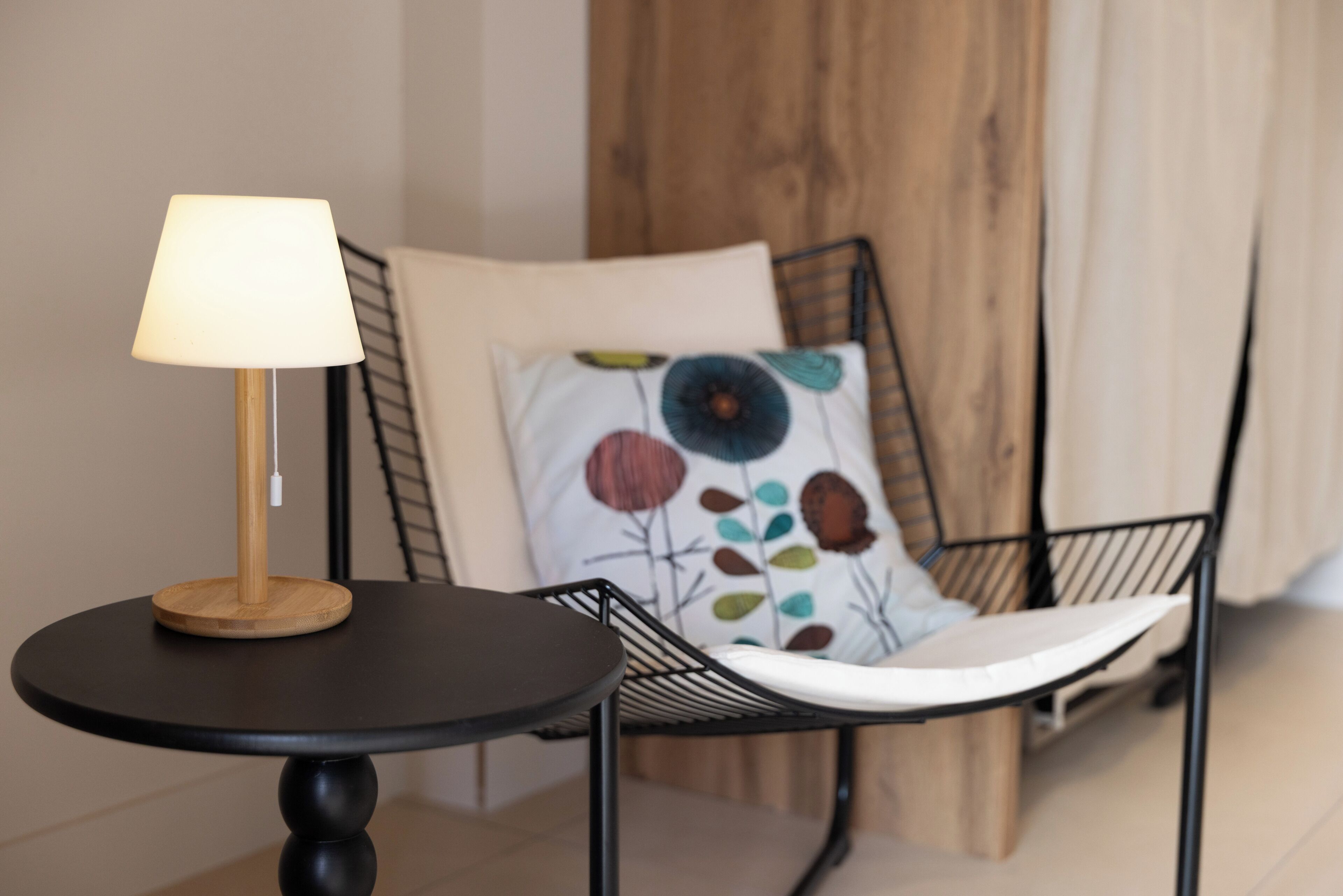 Une table ronde avec une lampe, une chaise en métal noir avec un coussin, et une chaise en bois avec un coussin à motifs floraux.
