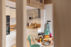 A modern kitchen with a dining area, featuring a wooden countertop, a built-in oven, and a dining table set with plates, fruits, and a baguette.