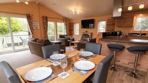 A dining area with a wooden table set for two, a kitchen in the background, and a living area with sofas and a TV. - L21 Coastal Kippford (Dalbeattie)