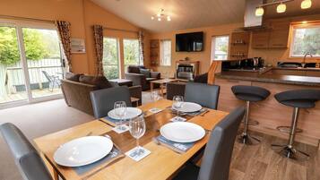 A dining area with a wooden table set for two, a kitchen in the background, and a living area with sofas and a TV.