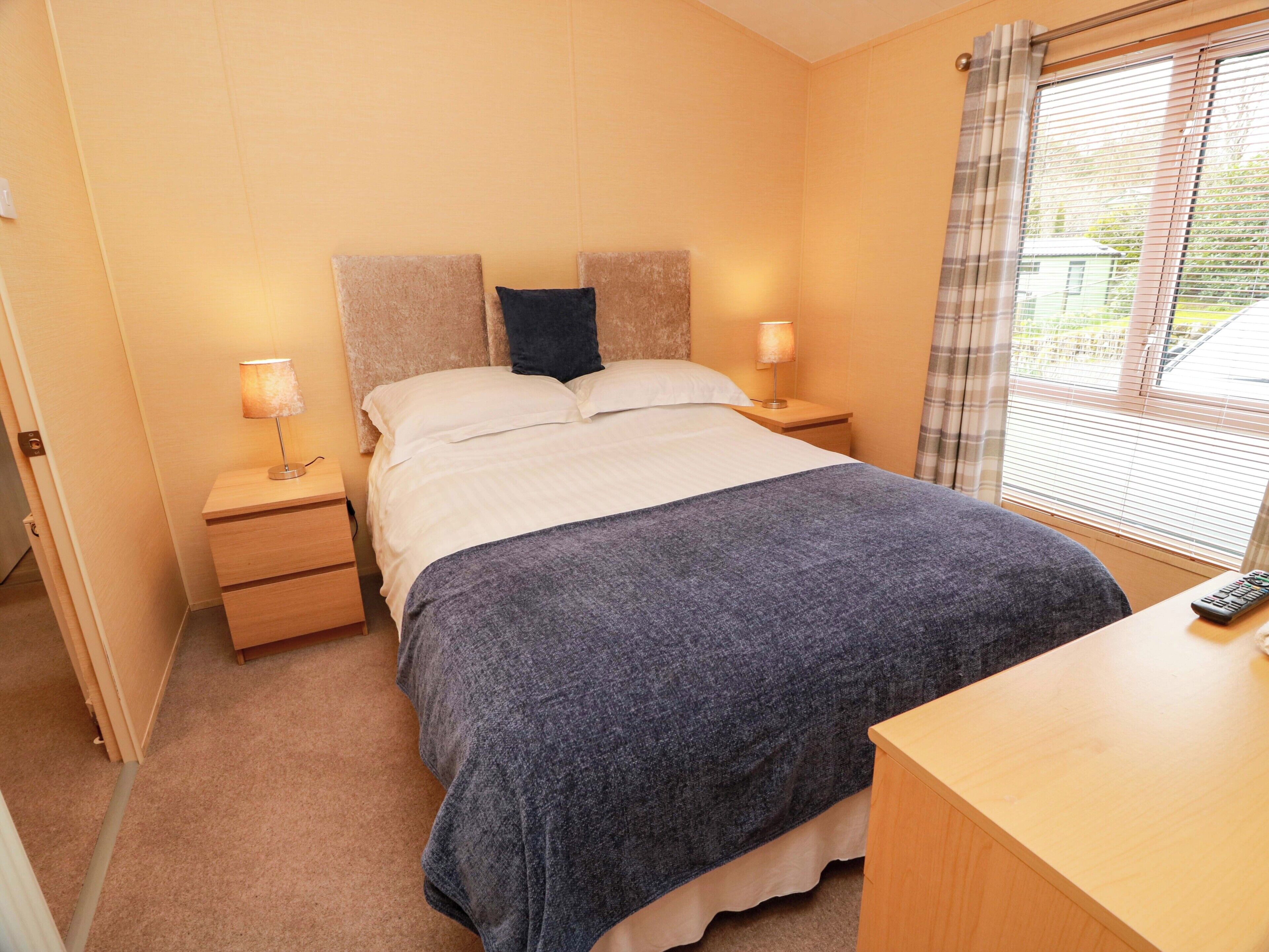 A bedroom with a bed, two bedside tables, a nightstand, a window with blinds, and a remote control on the nightstand.
