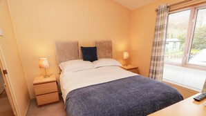 A bedroom with a bed, two bedside tables, a nightstand, a window with blinds, and a remote control on the nightstand. - L21 Coastal Kippford (Dalbeattie)