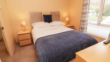 A bedroom with a bed, two bedside tables, a nightstand, a window with blinds, and a remote control on the nightstand.
