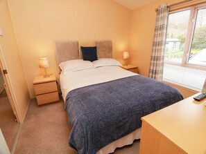 A bedroom with a bed, two bedside tables, a nightstand, a window with blinds, and a remote control on the nightstand. - L21 Coastal Kippford (Dalbeattie)
