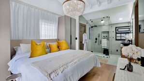 Ett modernt hotellrum med en stor säng, en kristallkrona, en dusch med glasväggar och ett badrum med spegel och handfat.