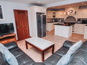 Ein modernes Wohnzimmer mit einer Ledersofa, einem Holztisch, einem Fernseher auf einem Holzständer sowie einer Küche mit weißen Schränken und einer schwarzen Arbeitsplatte.