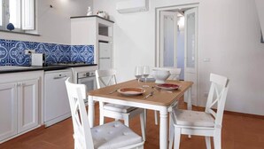 Een keuken met witte kastjes, een betegelde spatwand, een eettafel met stoelen en een raam met zonwering.