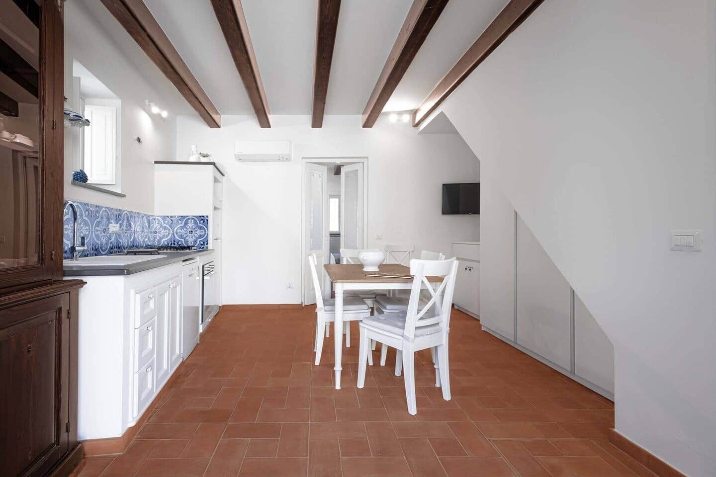 A kitchen with white cabinets, a blue patterned backsplash, a dining table with chairs, and a TV mounted on the wall.