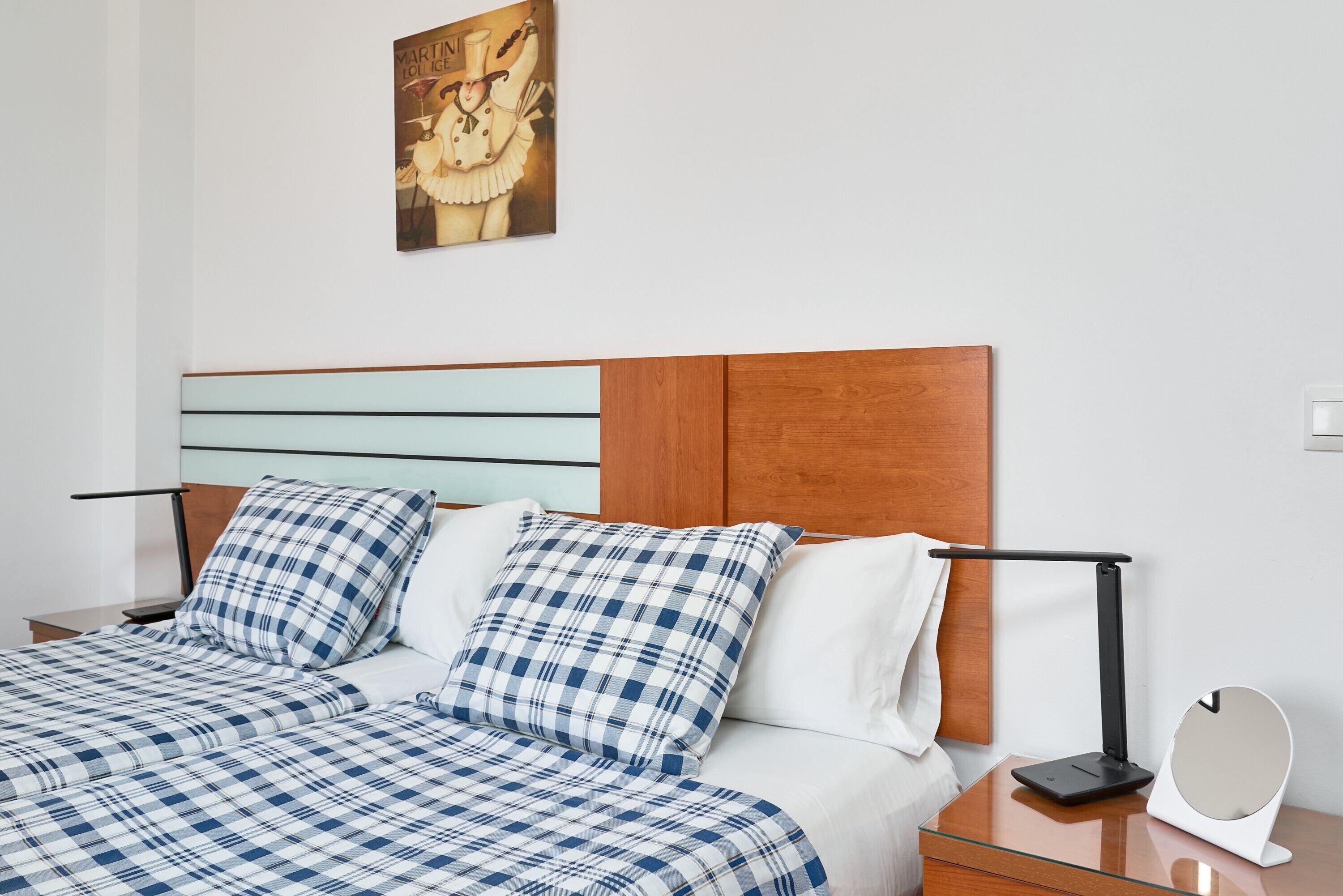 Una cama bien hecha con una colcha a cuadros, cabecera de madera, mesita de noche con lámpara y un cartel en la pared.