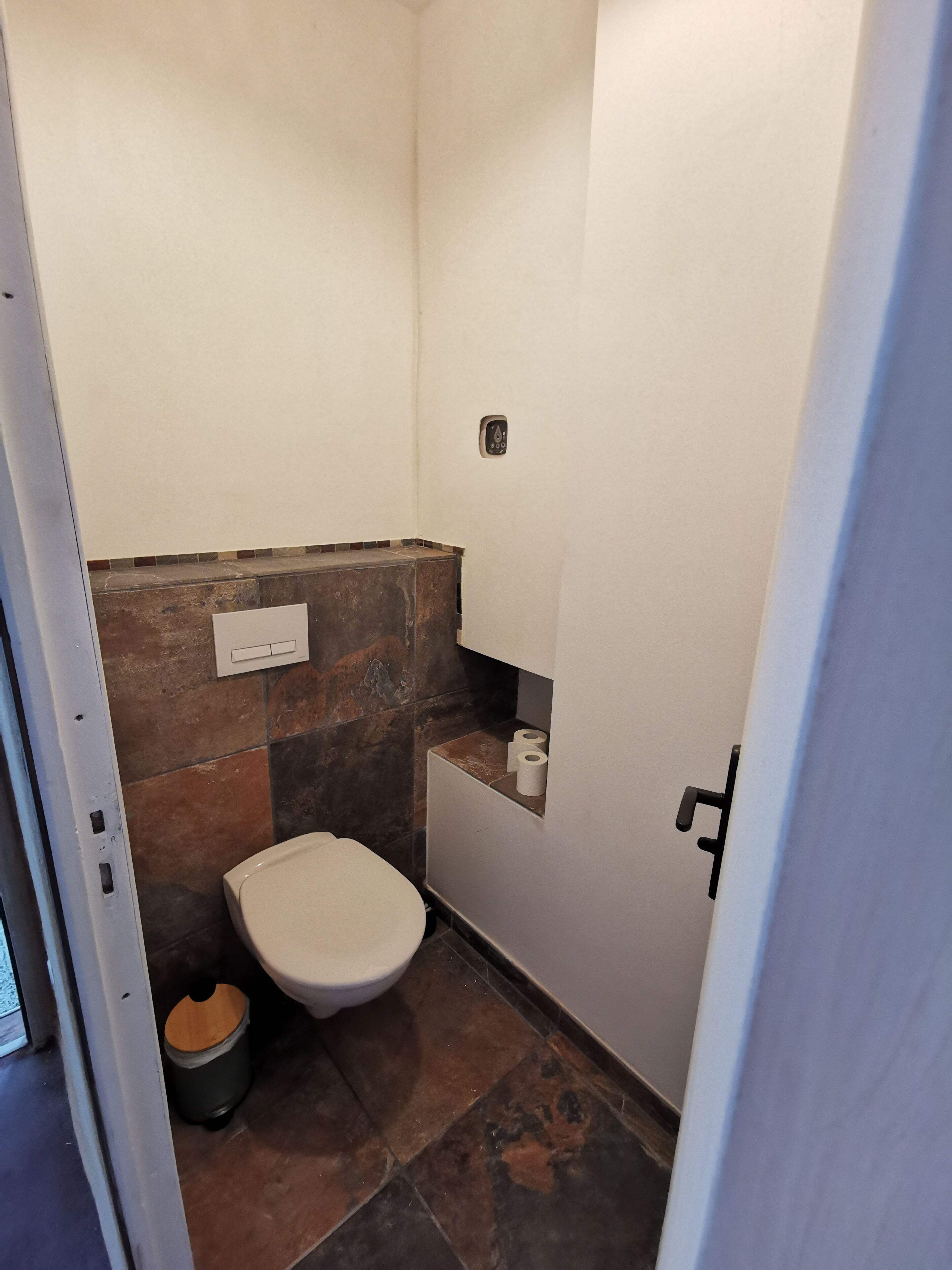 A small, compact bathroom with a toilet, a trash bin, and a shelf with toilet paper.
