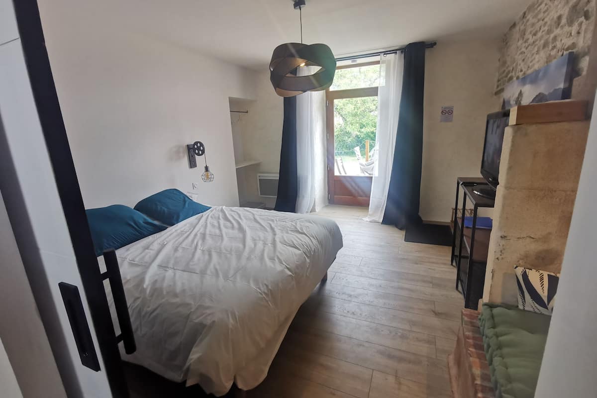 A bedroom with a bed, a desk, a chair, a hanging light fixture, and a view of the outdoors.