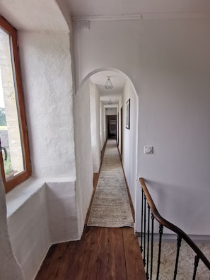Un pasillo con piso de madera, paredes blancas y una ventana a la izquierda.