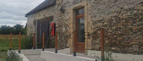 Un edificio de piedra con puerta de madera, una ventana pequeña y un camino de grava que lleva a un banco de madera.
