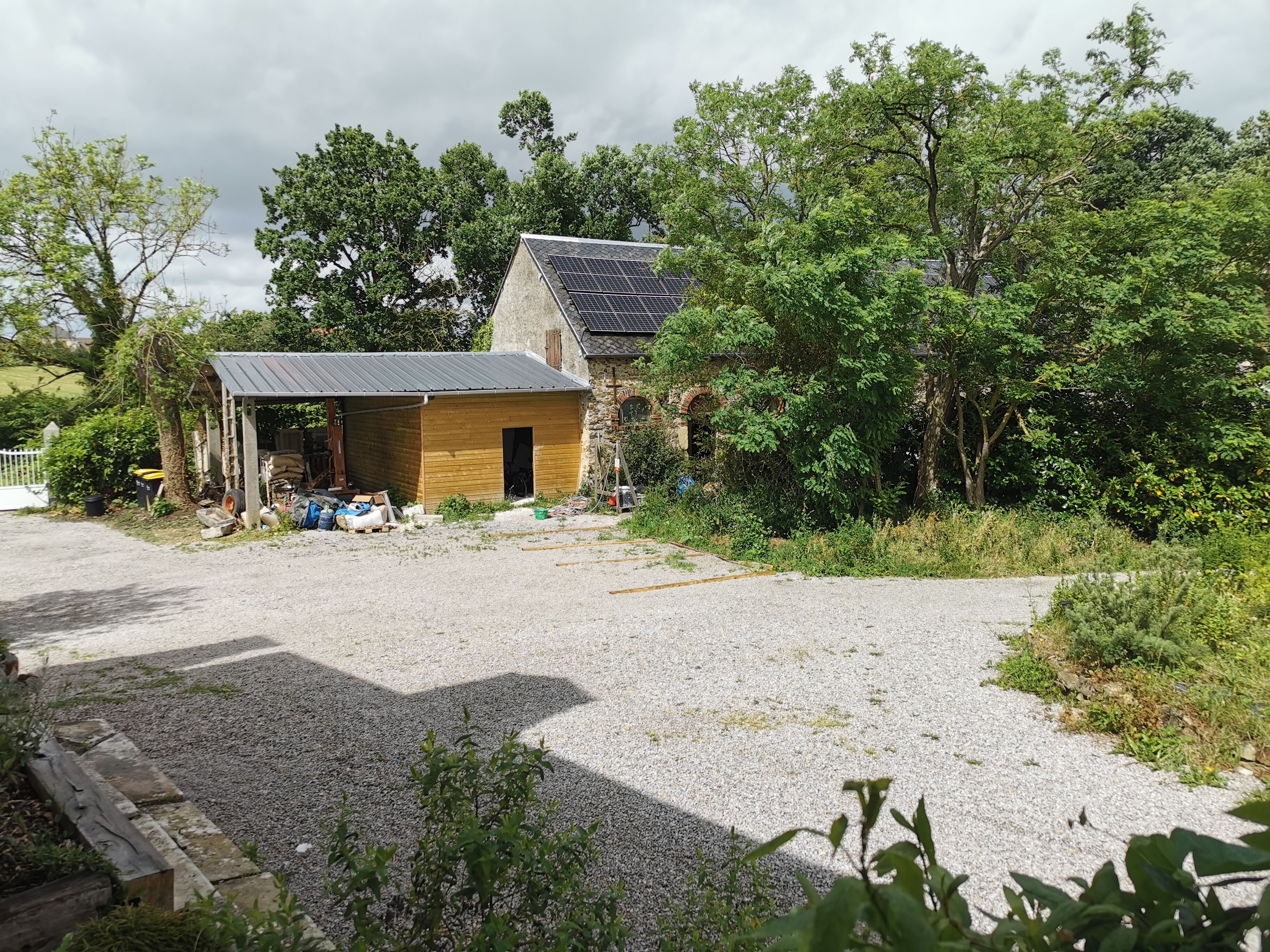 A gravel driveway leading to a wooden shed and a stone building with a solar panel roof.