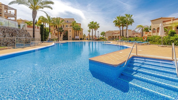 A swimming pool with clear blue water, surrounded by palm trees and hotel buildings. - Luxury 2BR Resort Apt, Pools, Spa, Tennis (Estepona)
