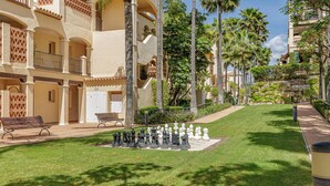 A courtyard with a chessboard, benches, palm trees, and a building with balconies. - Luxury 2BR Resort Apt, Pools, Spa, Tennis (Estepona)