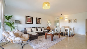 A living room with a sofa, a rocking chair, a coffee table, a dining area with chairs, and a chandelier. - Luxury 2BR Resort Apt, Pools, Spa, Tennis (Estepona)