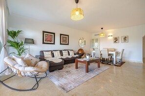 A living room with a sofa, a rocking chair, a coffee table, a dining area with chairs, and a chandelier. - Luxury 2BR Resort Apt, Pools, Spa, Tennis (Estepona)